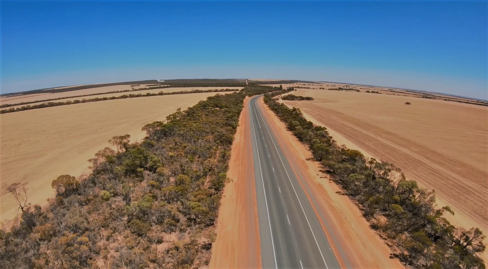 Aerial view of a sealed rural highway stretching to the horizon through golden Western Australian farmland and native bushland under a clear blue sky
