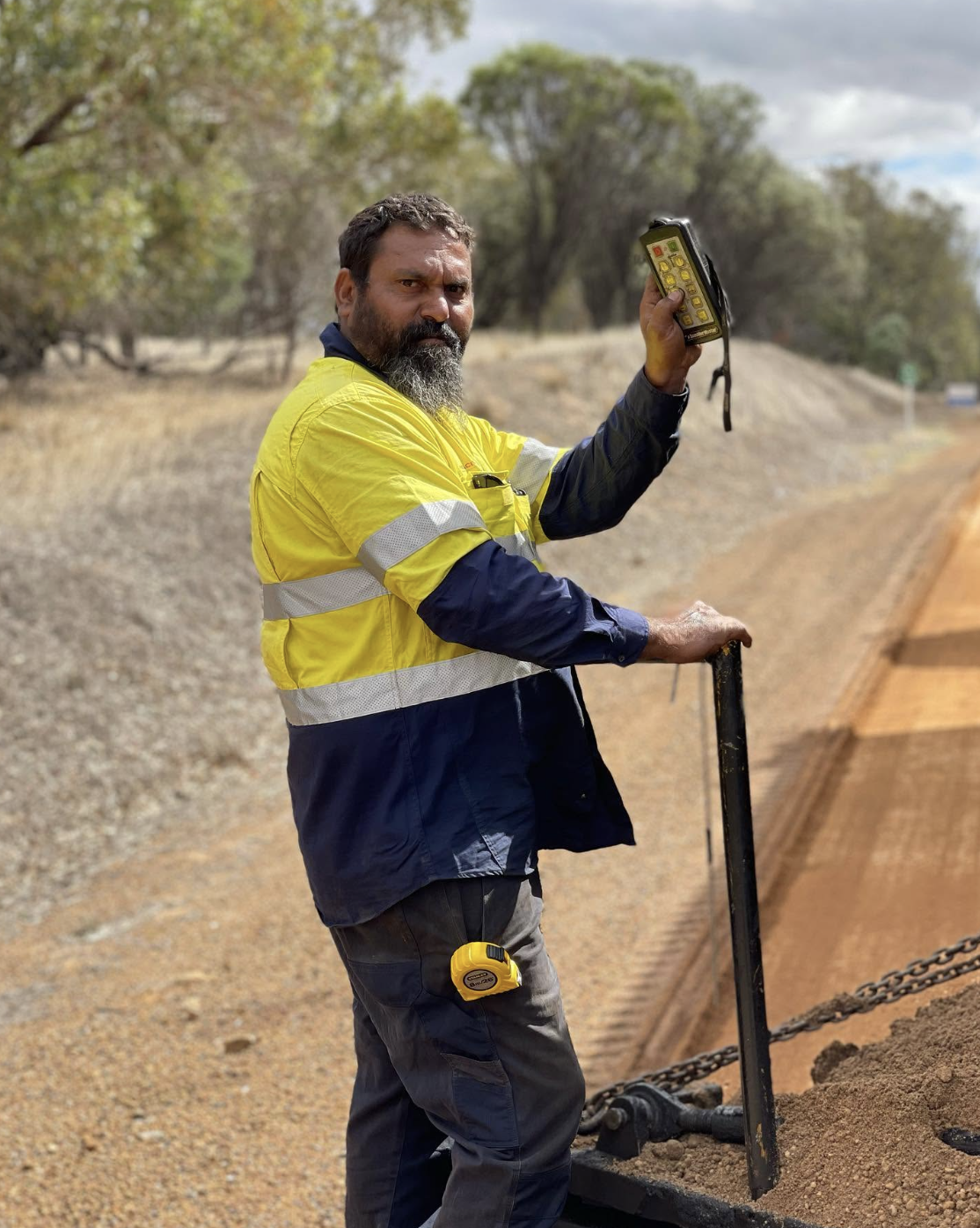 YongaCivil worker holding a remote control on the back of a truck spreading gravel along a rural road