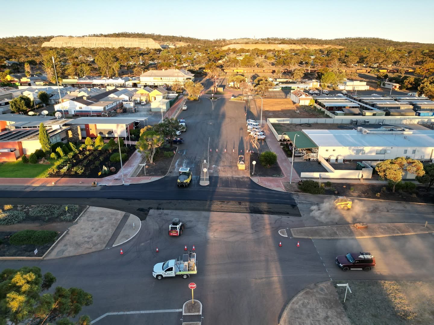 Aerial view of a regional Western Australian town intersection being resurfaced with fresh asphalt, a road roller, traffic management and YongaCivil crew at golden hour