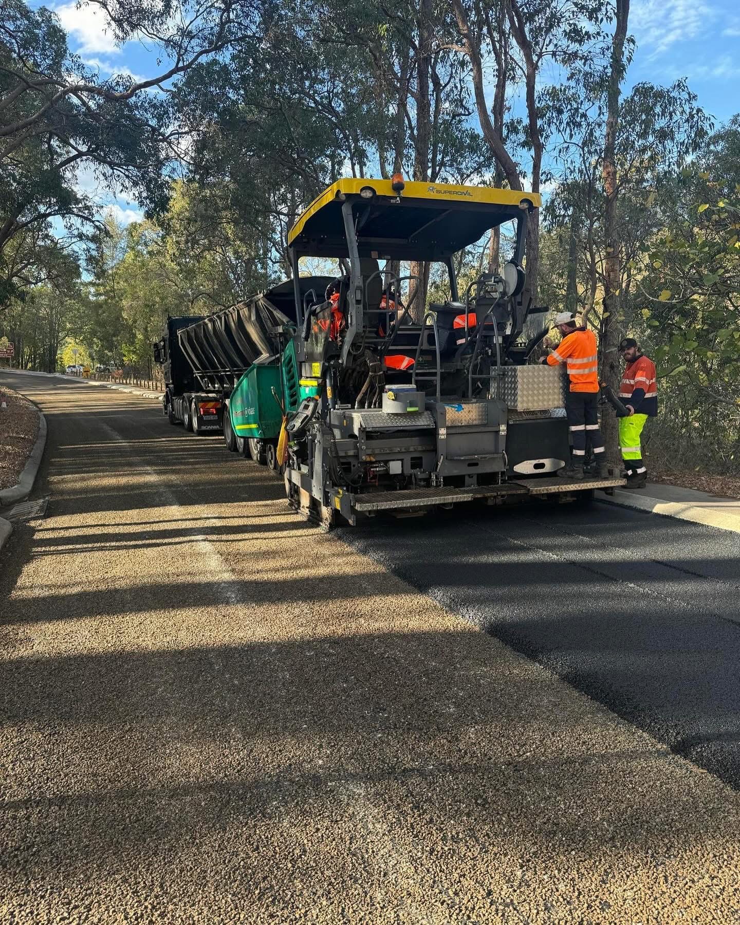 YongaCivil branded asphalt paver laying fresh asphalt on a tree-lined regional road with a Scania tipper truck feeding hot mix and hi-vis crew working