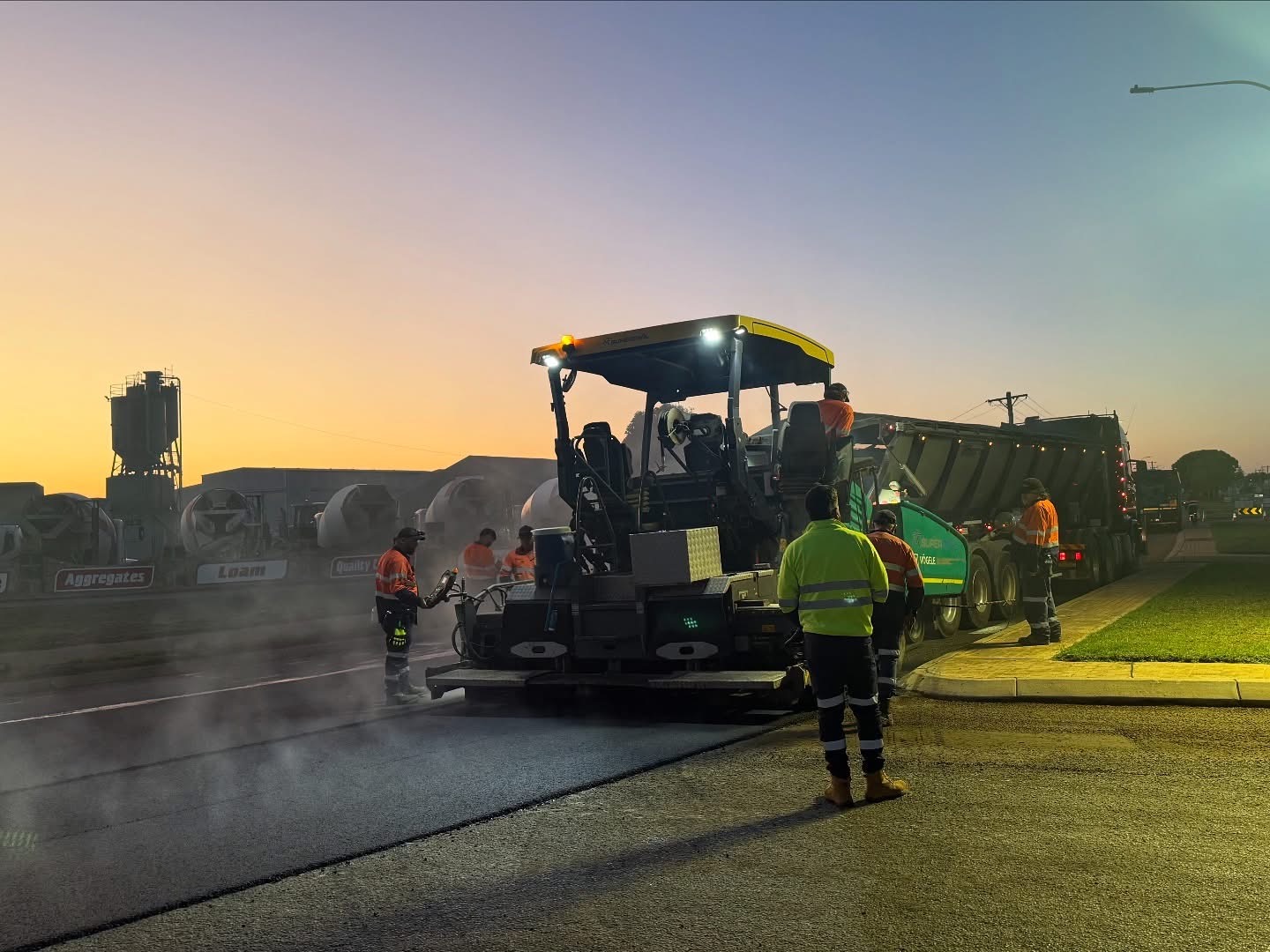 YongaCivil crew resurfacing a road at dawn with a Vögele paver laying fresh asphalt beside a tipper truck and aggregate supply yard