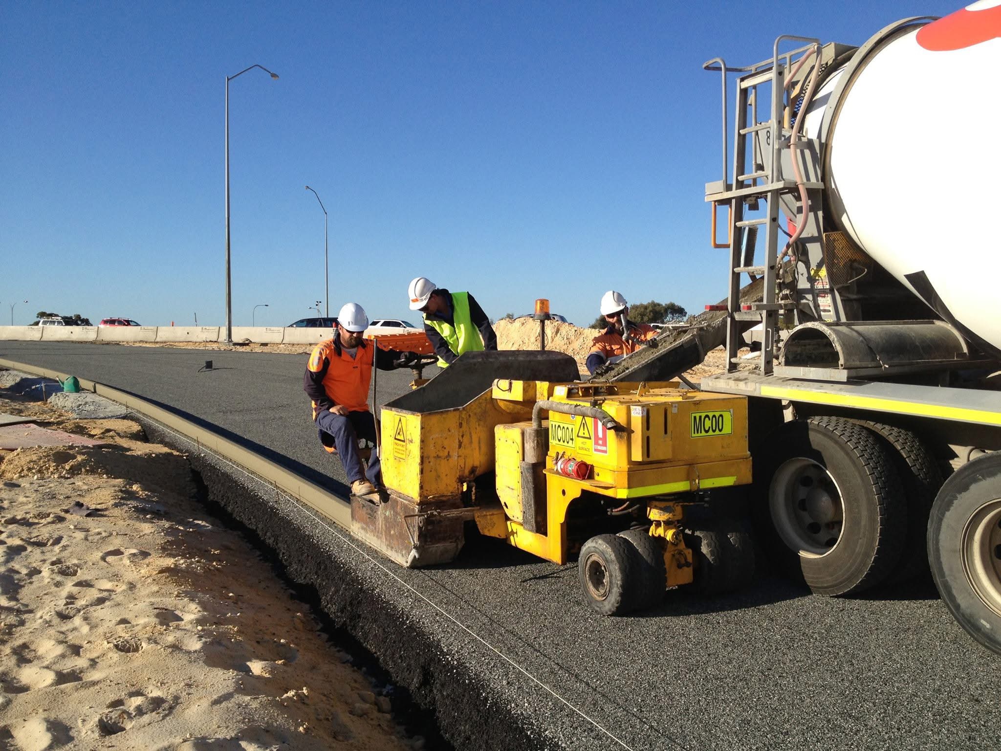 YongaCivil crew operating a slipform kerbing machine fed by a concrete agitator truck, extruding a fresh continuous concrete kerb along a newly paved Western Australian road