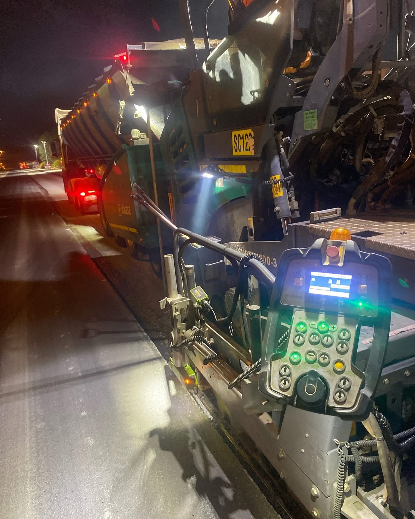 YongaCivil Vogele asphalt paver working under floodlights during night road construction works, with its illuminated control console in the foreground and a material feed truck ahead