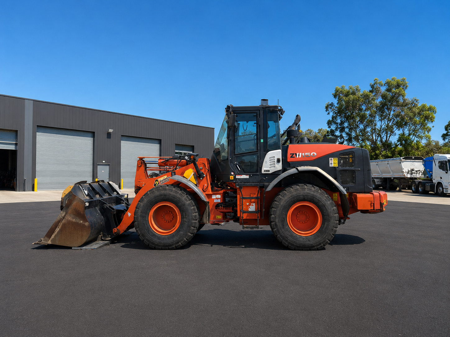 Orange Hitachi ZW150 IT loader with a front bucket, parked on asphalt outside an industrial warehouse under a clear blue sky in Western Australia