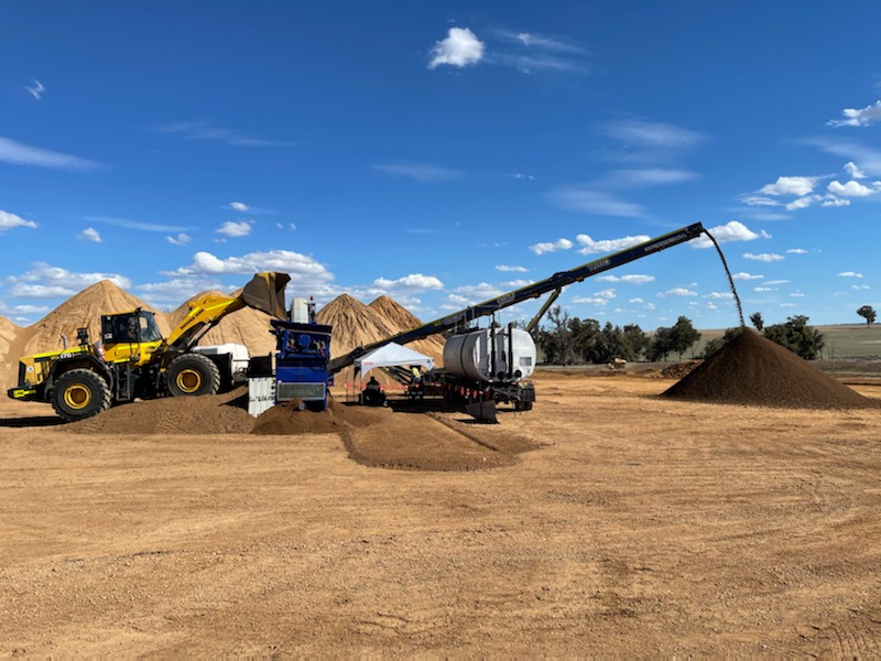 Blue YongaCivil pugmill plant blending stabilised material on a rural Western Australian site, with a wheel loader feeding stockpiled material and the conveyor discharging into a large stockpile under a blue sky