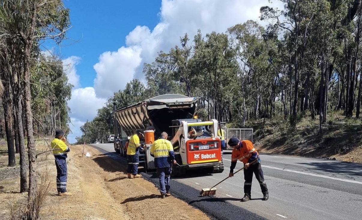 Hi-vis YongaCivil crew carrying out shoulder construction on a regional Western Australian highway with a Bobcat skid steer and tipper truck laying fresh asphalt while workers rake and sweep the new road shoulder