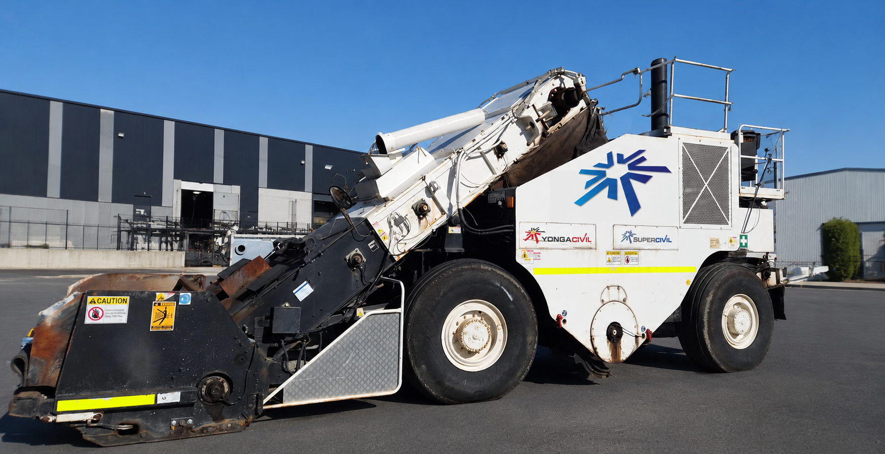 White YongaCivil shuttle buggy material transfer vehicle with raised conveyor arm, parked outside an industrial warehouse under a clear blue sky in Western Australia
