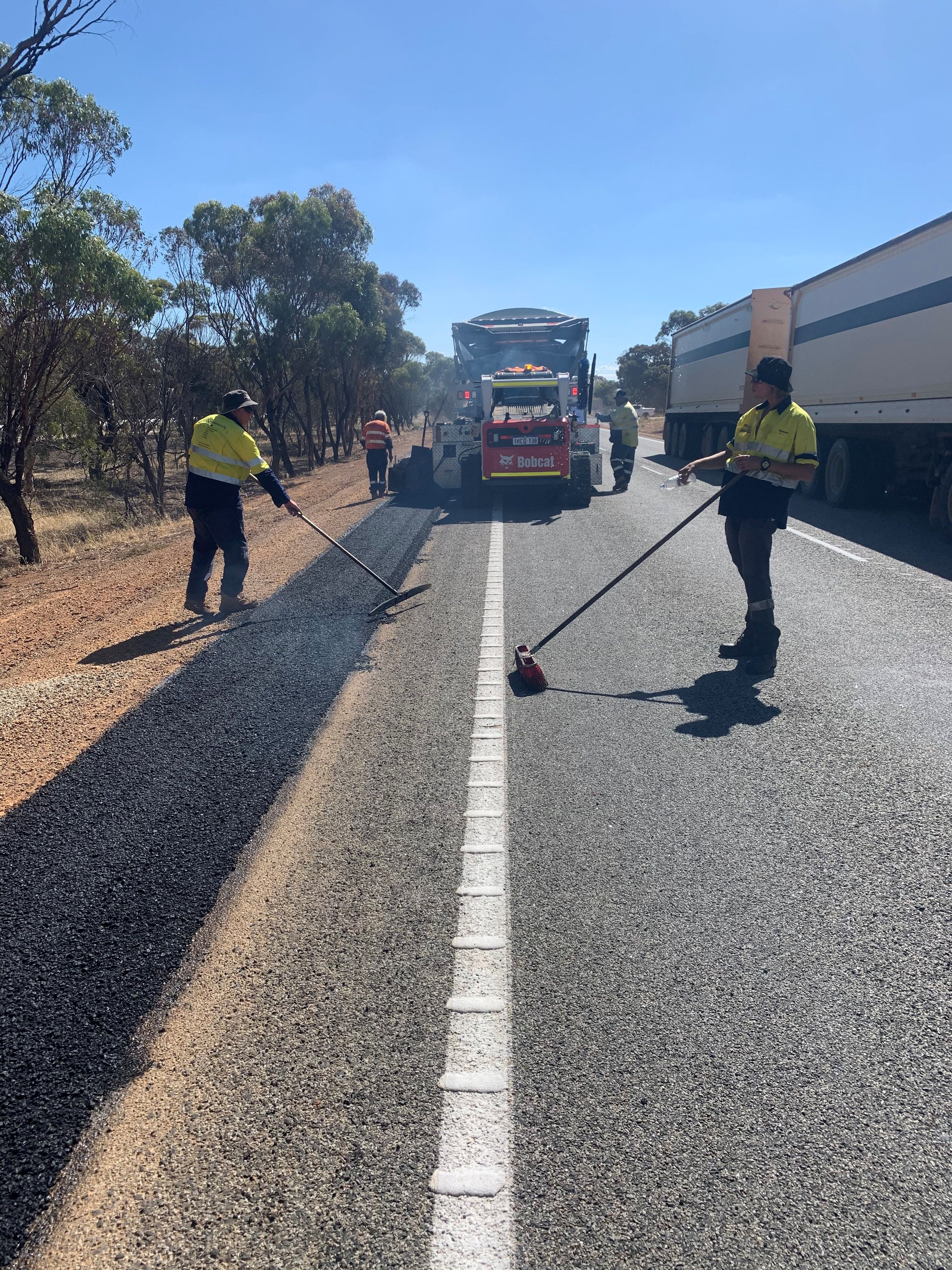 YongaCivil crew in hi-vis raking and spreading fresh asphalt to widen the shoulder of a regional Western Australian highway with a Bobcat skid-steer feeding material