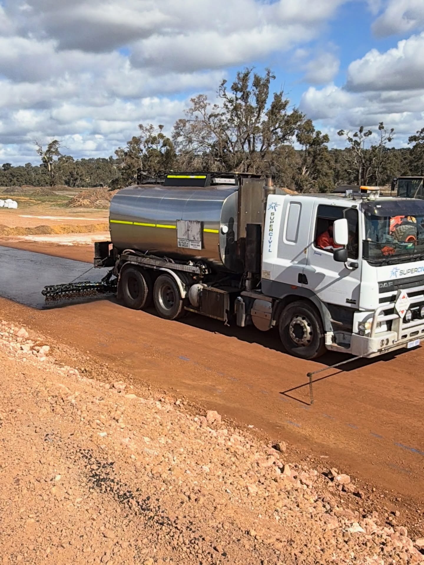 YongaCivil-branded DAF tanker truck with a rear spray bar applying water to a compacted red-dirt road base on a regional Western Australian construction site