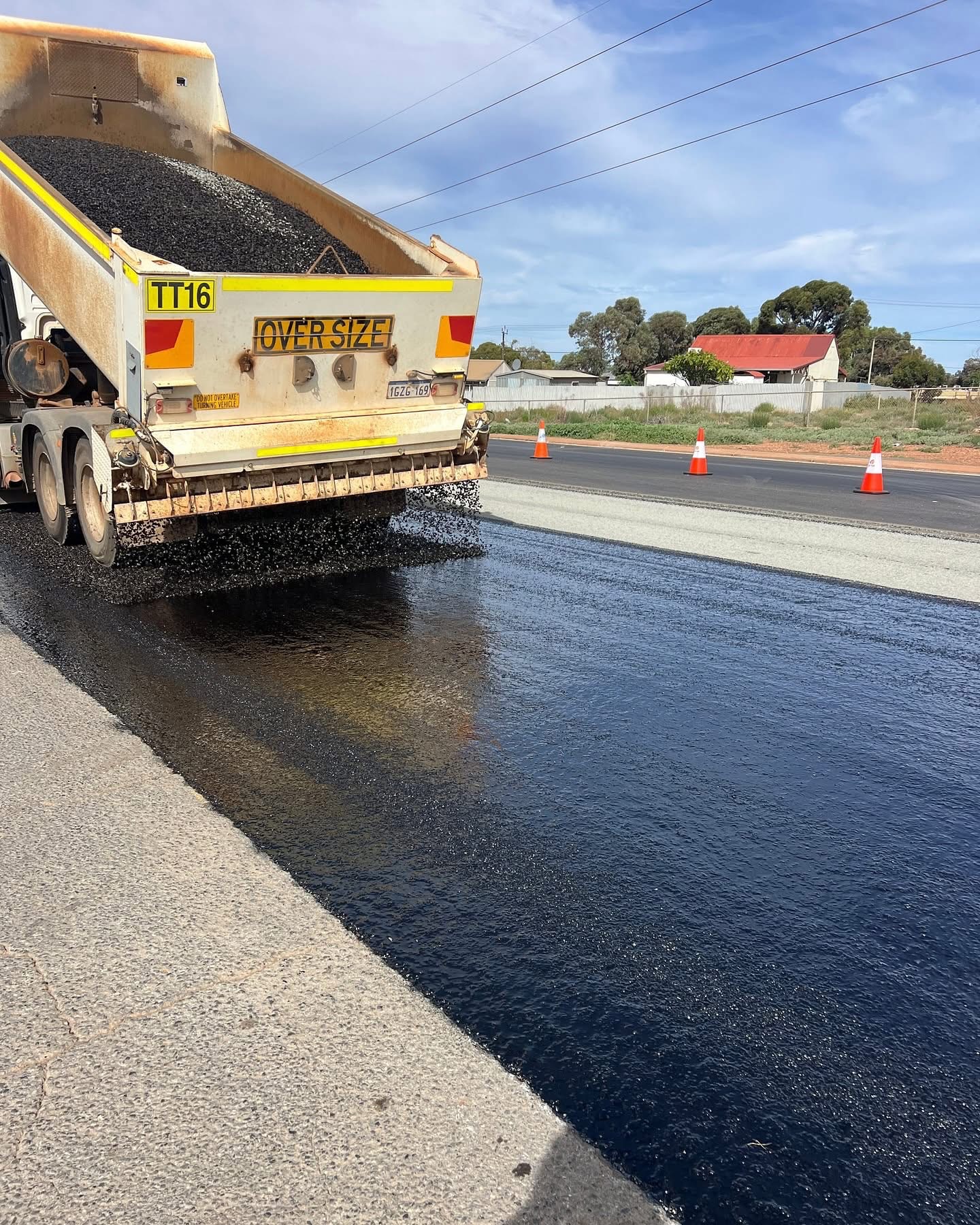 Oversize aggregate spreader truck discharging aggregate over a freshly spray-sealed bitumen strip on a regional Western Australian road, marked with traffic cones