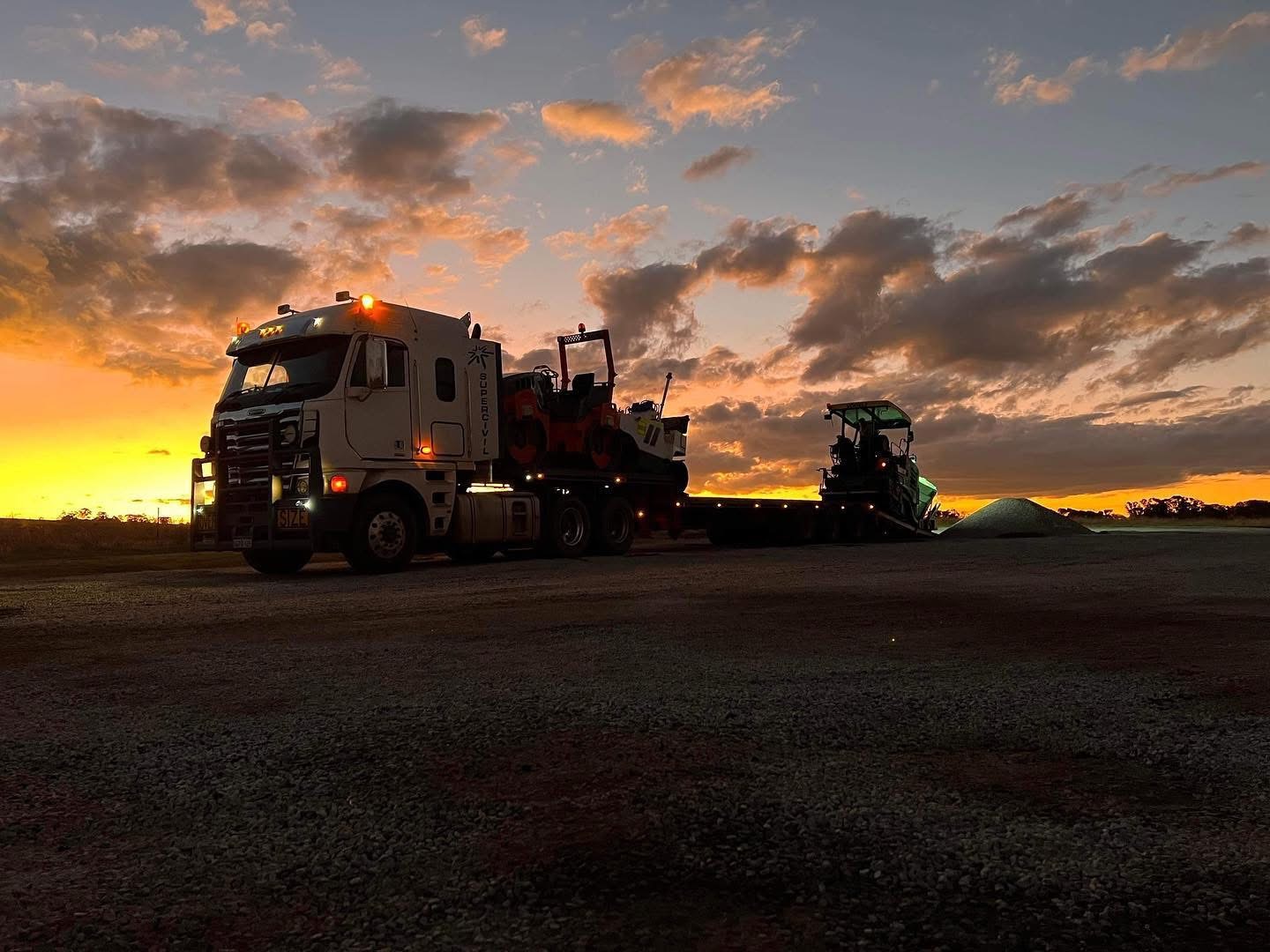 White YongaCivil prime mover with a low-loader float carrying a compaction roller and civil plant, parked on a gravel pad against a golden sunset in regional Western Australia