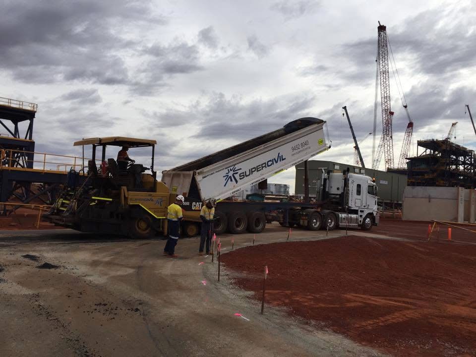 YongaCivil semi end tipper with its trailer raised discharging asphalt into a Caterpillar paver on a red-dirt mine site, with a hi-vis crew and cranes in the background