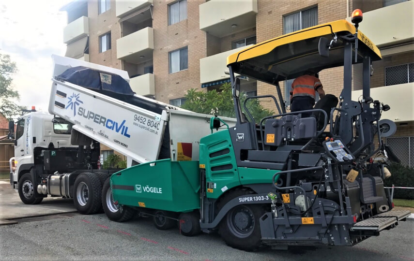 YongaCivil 6-wheel tipper with its body raised discharging asphalt into a green Vögele paver on a suburban street in front of a residential apartment block