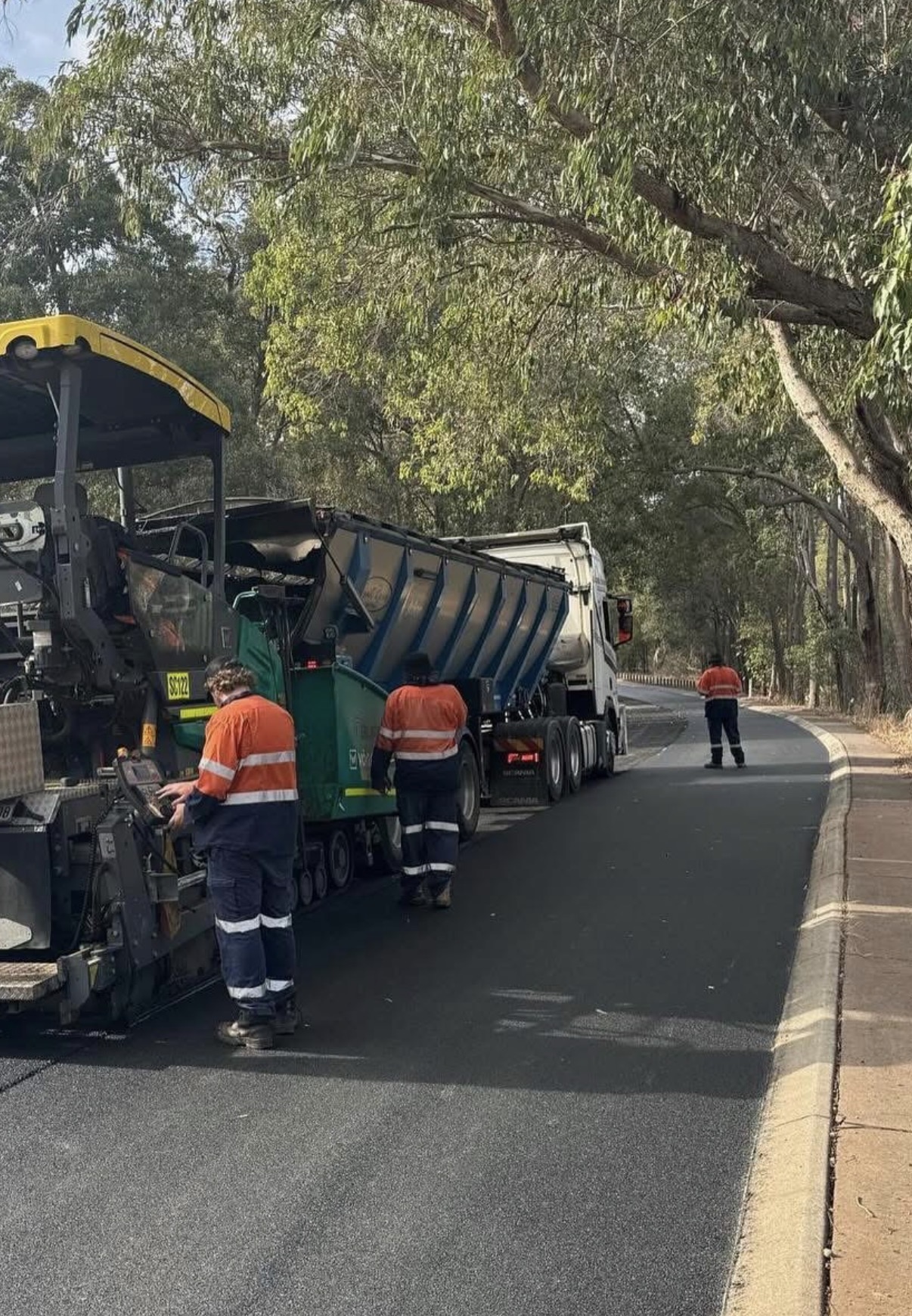 YongaCivil walking floor trailer feeding asphalt into a paver on a tree-lined suburban Perth road while a hi-vis crew lays a fresh asphalt mat